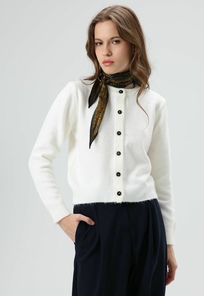 Cardigan blanc en tricot avec boutons noirs, encolure ronde et poignets côtelés. Porté sur un pantalon bleu marine, complété par une écharpe noire à motifs dorés.