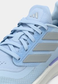 Αθλητικό παπούτσι ανοιχτού γαλάζιου χρώματος με υφασμάτινο επάνω μέρος με υφή, επίπεδα κορδόνια και γκρι λεπτομέρειες, διαθέτοντας το λογότυπο με τρεις ρίγες στην πλευρά.