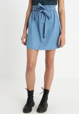 Kobieta ubrana w jasnoniebieską spódnicę dżinsową z paskiem wiązanym w talii oraz czarne, sznurowane botki za kostkę, stojąca na tle jednolitego tła.