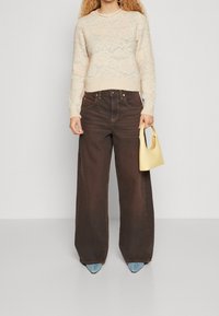 Beiger pullover mit Spitzenmuster, braune Jeans mit weitem Bein, blaue spitze High Heels und eine kleine gelbe Handtasche. Strukturierter Strick und hoher Bund.