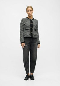 Schwarze und weiße Tweedjacke mit rundem Hals, drei Metallverschlüssen, zwei Fronttaschen und schwarzem Besatz. Kombiniert mit grauen Jeans und schwarzen Loafers.