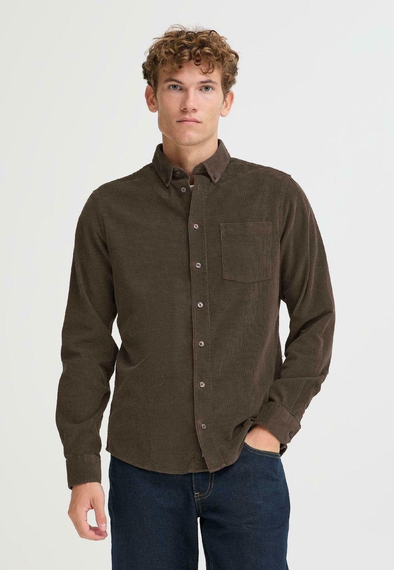 Camisa de pana marrón oscuro con cuello abotonado, un bolsillo en el pecho y cierres de botones, combinada con jeans azules. Diseño texturizado y estructurado.