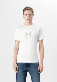 Hvit bomull t-skjorte med korte ermer, med et stort, lys gul "H" logo og ordet "HACKETT" plassert under. Kombinert med blå jeans.