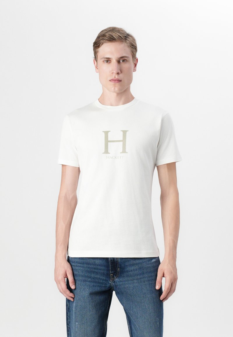 Hvit bomull t-skjorte med korte ermer, med et stort, lys gul "H" logo og ordet "HACKETT" plassert under. Kombinert med blå jeans.