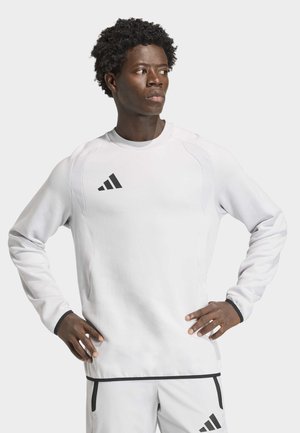 Mężczyzna ubrany w biały dres Adidas z czarnym logo, stoi z rękami na biodrach, patrząc na bok na białym tle.