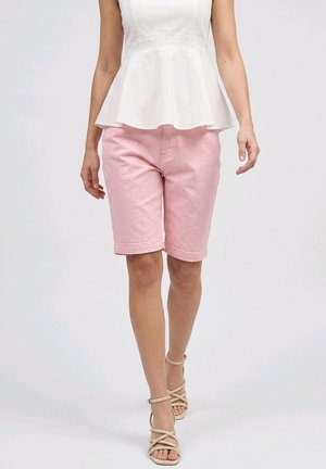 Mujer con top blanco sin mangas con peplum, pantalones cortos rosa hasta la rodilla y sandalias beige con tiras, de pie contra un fondo liso.