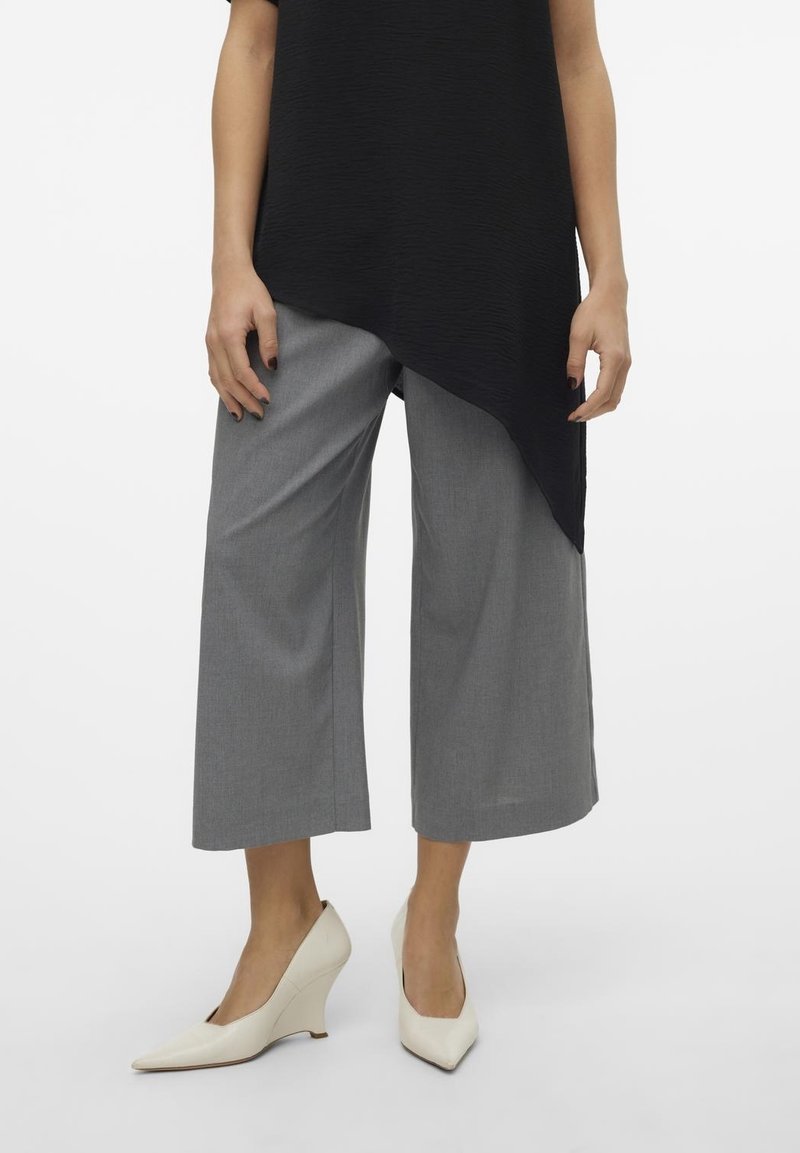 Femme portant un pantalon gris large et court, des chaussures compensées crème à bout pointu et un haut asymétrique noir, debout sur un fond blanc.