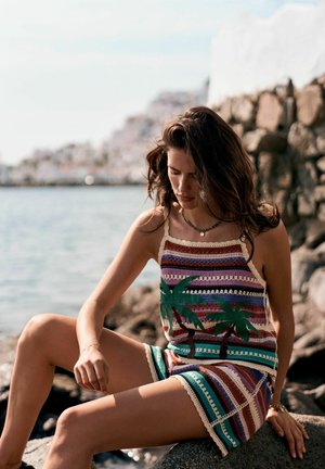 Ung kvinde i farverig stribet hæklet tanktop og shorts med palmetræsmønstre sidder på klipper ved vandet nær en stenmursmur.