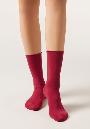 Jambes portant des chaussettes rouges unies mi-mollet, debout sur un fond clair uni.