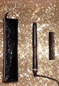 Schwarzes, strukturiertes Aufbewahrungstäschchen, schwarzes zylindrisches Stylinggerät mit einem Netzschalter und ein schwarzer Kamm auf einem goldglitzernden Hintergrund.