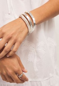 Three silver cuff bracelets with textured patterns stacked on a wrist, featuring a smooth, shiny finish and a matching silver ring.