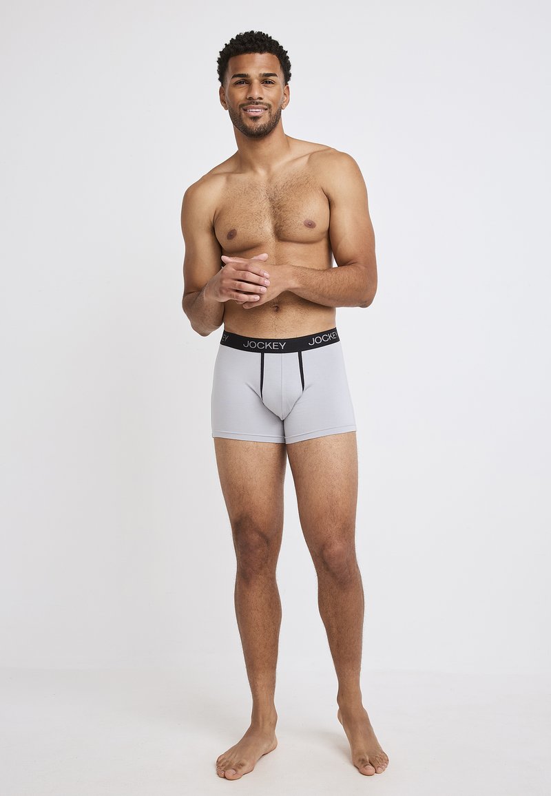 Gray boxer shorts made of smooth fabric, featuring a black branded waistband and a fitted design. Model stands barefoot, arms relaxed.