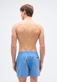 Ανοιχτό μπλε μαγιό σορτς με ελαστική μέση, πίσω τσέπη και λεία υφή, που φοριέται από έναν άνθρωπο εμφανιζόμενο από πίσω.