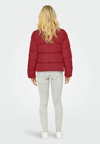 Rød pufferjakke med vandrette quiltede sektioner, parret med lysegrå slim-fit jeans og hvide sneakers, set fra ryggen.