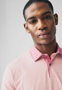 Rosa pikétröja med en mönstrad krage i en mörkare nyans av rosa, med kontrasterande knappar och en slät textur.