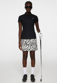 Czarna, teksturowana koszulka polo z kołnierzykiem, zestawiona z białą spódnicą w czarny wzór zebry. Czarne buty i rękawiczka z kijem golfowym.