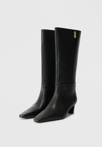 Schwarze Leder-Kniestrumpf-Stiefel mit spitz zulaufender Zehe und einem niedrigen Blockabsatz, der an der Seite mit einem goldfarbenen Akzent verziert ist. Glatte Textur und elegantes Design.