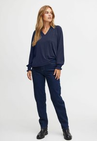 Mörkblå V-ringad topp med långa, något puffiga ärmar; lätt material med rynkade detaljer vid ärmsluten. Mörka jeans och svarta ankelstövlar.
