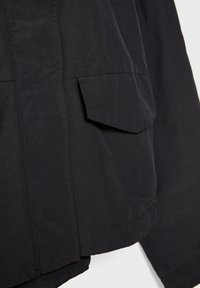 Gros plan d'une poche de manteau noire avec rabat et coutures visibles sur un tissu lisse.