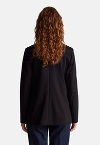 Černý blazer s strukturovaným designem, hladkým materiálem a švy s kroucením vzadu. Modelka má kudrnaté vlasy a nosí džíny.