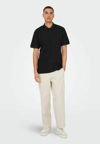 Sort kortærmet polo-shirt med krave, båret med lysebeige bukser og hvide sneakers. Glat stof, afslappet pasform.