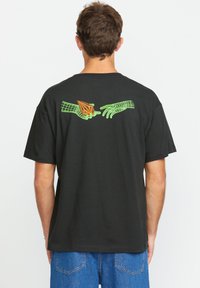 Schwarzes T-Shirt mit kurzen Ärmeln, das auf der Rückseite ein Grafikdesign mit grünen Händen und einem orangefarbenen Objekt zeigt. Aus Baumwolle, lässiger Schnitt.