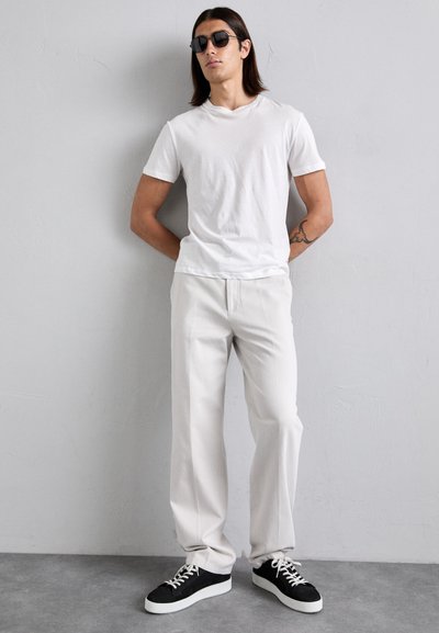 Young man with medium-length hair wearing black sunglasses, white t-shirt, light pants, and black sneakers, leaning against a gray wall.