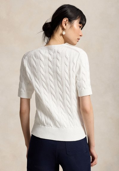 Kobieta z ciemnymi włosami związanymi w kok, ubrana w biały, krótki sweter z warkoczowym splotem i ciemne spodnie, zwrócona bokiem, prezentująca tył swetra.