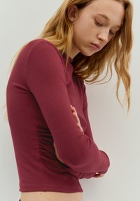 Dolga majica z dolgimi rokavi, prilegan kroj, v globoki vinsko rdeči barvi, iz bombaža; ima naguban detajl ob straneh, okrogel izrez in gladko teksturo.
