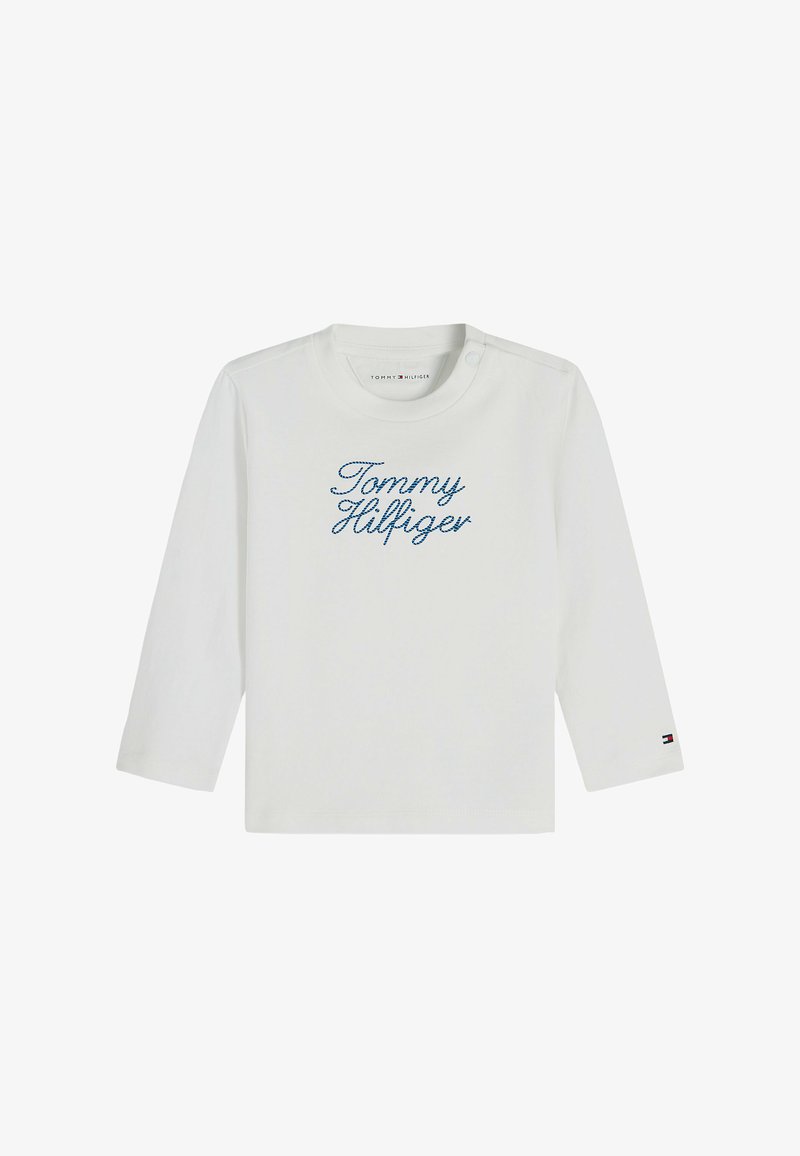Chemise blanche à manches longues avec « Tommy Hilfiger » en script bleu sur le devant et un petit logo Tommy Hilfiger sur le poignet de la manche gauche.