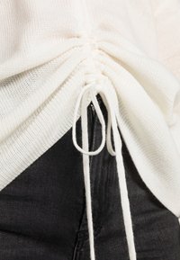 Weißes gestricktes Oberteil mit gerafften Seiten und einem Kordelzugdetail. Kombiniert mit schwarzem Denim, der Textur und kontrastierende Farben zur Schau stellt.