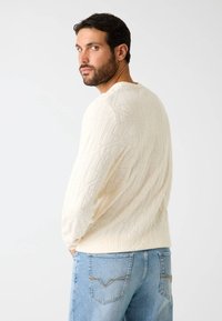 Jasnokremowy sweter dzianinowy z teksturowanym geometrycznym wzorem, okrągłym dekoltem i długimi rękawami, zestawiony z jasnoniebieskimi dżinsami.