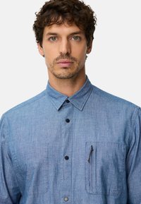 Hellblaues Chambray-Hemd mit Knopfkragen, Brusttasche vorne und einem Reißverschlusstaschen-Detail. Es hat dunkle Knöpfe und eine dezente Struktur.