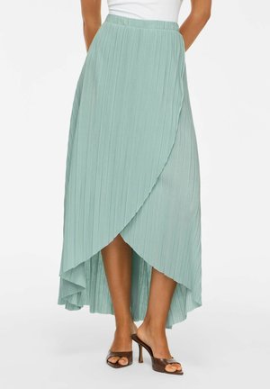 Femme portant une jupe portefeuille plissée vert clair avec un ourlet asymétrique, associée à des chaussures à talons hauts ouvertes marron.