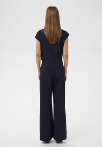 Salopetă marină cu mâneci scurte tip capac, talie ajustată, pantaloni cu picioare largi și detaliu la buzunarul din spate. Țesătură netedă, cu textură minimă.