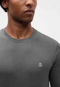 T-shirt grigio a maniche lunghe realizzata in tessuto morbido, con scollatura rotonda e un piccolo logo bianco sul petto. Texture liscia e design aderente.