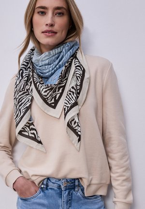 Giovane donna che indossa un maglione beige, jeans di denim blu e sciarpe sovrapposte con stampa zebra e motivo blu intorno al collo, in piedi contro una parete liscia.