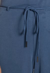 Blå tygbyxor med dragsko. Dragsnörena har svarta metallspetsar. Materialet har en mjuk, matt yta.