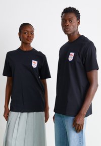 Melna kokvilnas T-kreklu ar īsām piedurknēm, uz kura redzams izcils sarkans un zils Adidas logotips uz vairoga emblemas, ko valkā divi modeļi.