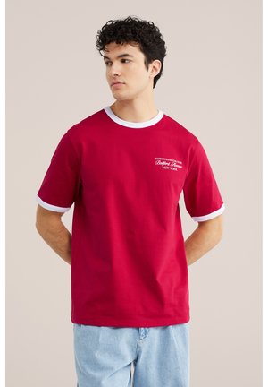 Ung mand iført en rød T-shirt med hvide kanter og lyseblå jeans, der kigger til siden med hænderne bag ryggen.