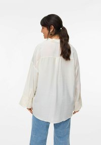 Camicia oversize di un cremoso chiaro con maniche lunghe, orlo arrotondato e tessuto testurizzato. Abbinata a pantaloni in denim blu chiaro.
