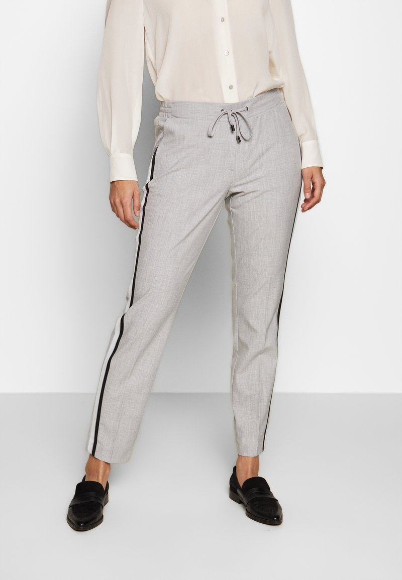 Femme portant un pantalon gris clair sur mesure avec des bandes latérales noires et un cordon de serrage, associé à un chemisier crème et des mocassins noirs.