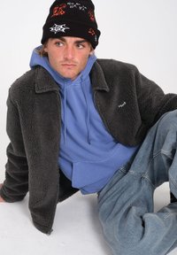 Giacca in pelliccia sintetica grigia con colletto, indossata sopra una felpa con cappuccio azzurra. Beanie nero lavorato a maglia con motivi grafici. Jeans in denim azzurro.