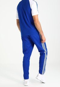 Sininen urheiluva Outfit, joka koostuu sinisestä lyhythihaisesta paidasta ja yhteensopivista juoksuhousuista, joissa on valkoiset sivuvoimat. Kankaan pinta näyttää sileältä; kengät ovat valkoiset.