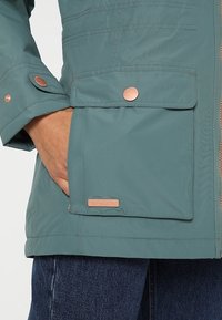 Main dans la poche d'une veste bleu sarcelle avec une poche avant à rabat et un bouton-pression en cuivre, associée à un jean bleu.