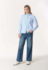 Donna in piedi con una mano in tasca, indossa una camicia a righe bianche e blu con bottoni, jeans a gamba larga e sneakers bianche su sfondo bianco.