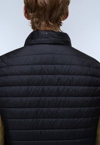 Rövid világosbarna hajú személy fekete steppelt dzsekiben, hátulnézetben, egyszínű világos háttér előtt.