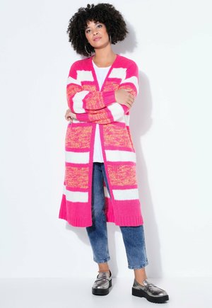 Kobieta o kręconych włosach stoi z założonymi skrzyżowanymi ramionami, ubrana w długi, różowo-biały kardigan w paski, białą koszulę, niebieskie dżinsy i metaliczne sandały na platformie.