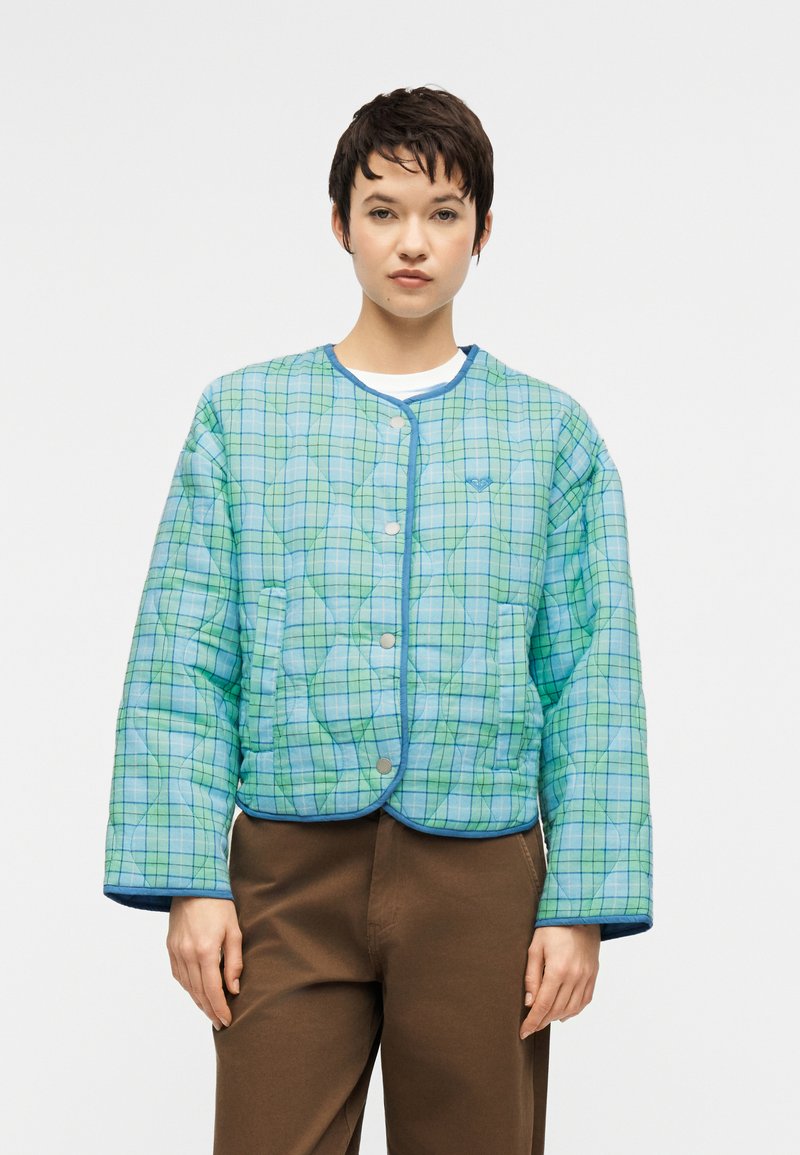 Γυναίκα με κοντά σκούρα μαλλιά φοράει μπλε και πράσινο καρό μπουφάν με ραφές και κουμπιά τύπου σναπ, και καφέ παντελόνι, στέκεται μπροστά σε απλό φόντο.