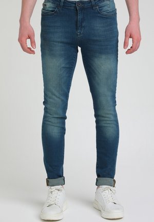 Mand iført tætsiddende blå jeans med opkrogede manchetter og hvide sneakers, stående mod en ensfarvet baggrund.